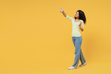 Fototapeta na wymiar Full size body length overjoyed african american young brunette woman 20s wears green shirt go walk do selfie shot on mobile cell phone show victory sign isolated on yellow background studio portrait.