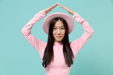 Happy charismatic young brunette asian woman 20s wears pink clothes hat hold folded hands above head like house roof isolated on pastel blue color background studio portrait. People emotions concept