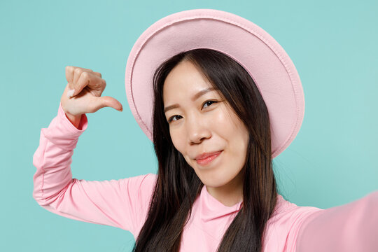 Close up young brunette asian woman 20s wears pink clothes wide brim hat doing selfie shot on mobile cell phone point thumb finger on herself isolated on pastel blue color background studio portrait