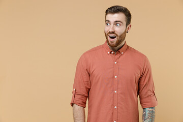 Obraz premium Surprised shocked tatooed young brunet man 20s short haircut with earrings wears apricot shirt look aside with open mouth isolated on pastel orange background studio portrait. People emotions concept.