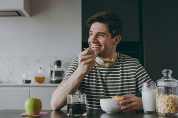 Young happy man in casual clothes striped t-shirt eat breakfast muesli cereals with milk fruit in bowl sit by table look aside cooking food in light kitchen at home alone Health diet lifestyle concept