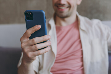 Young smiling satisfied happy man 20s wearing casual clothes beige shirt pink t-shirt use mobile cell phone in blue case sitting on grey sofa rest indoors at home on weekends. Focus on device gadget.