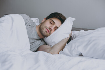 Fototapeta na wymiar Calm brunet tranquil caucasian young man 20s wearing pajamas grey t-shirt lying in bed sleep slumber resting with closed eyes relax at home indoors bedroom. Good mood night morning bedtime concept