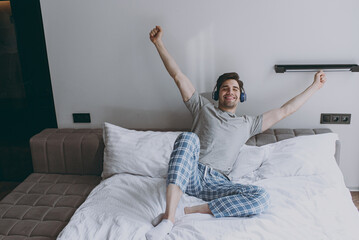Full size fun happy young man wear grey t-shirt blue pajamas headphones sit in bed listen to music do stretching rest at home on weekends relax indoors bedroom Good mood night morning bedtime concept.