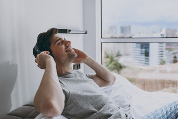 Side view happy relax satisfied young man wear grey t-shirt blue pajamas bottom headphones sit in...