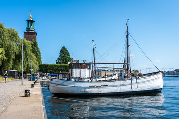 An old wooden fishing ship seiner is moored at the Stockholm waterfront. The City hall building is on the left. Panorama of the Swedish capital.