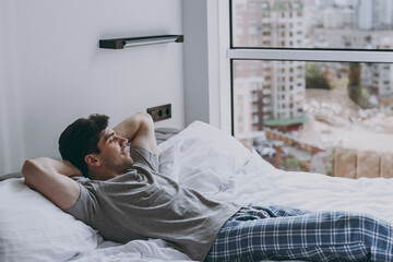 Side viw calm peaceful minded young man 20s wearing pajamas grey t-shirt lying in bed sleep slumber resting look at window relaxing at home indoors bedroom. Good mood night morning bedtime concept