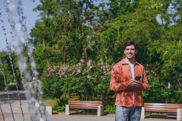 Fototapeta premium Young smiling fun man in orange jacket blue t-shirt walk using mobile cell phone near fountain rest relax in spring green city park go down alley sunshine lawn outdoor on nature Urban leisure concept