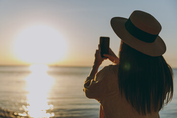Back view young woman in straw hat shirt summer casual clothes take photo of sunset by mobile cell phone resting outdoors at sunrise sun dawn over sea beach. People vacation lifestyle journey concept.