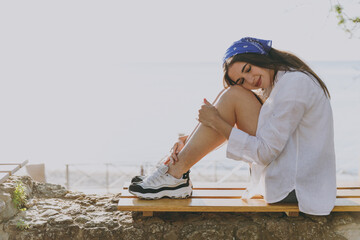 Side view full length young fun happy traveler tourist woman 20s in blue bandana shirt summer casual clothes rest sit on wooden bench outdoors at sea beach People vacation lifestyle journey concept.