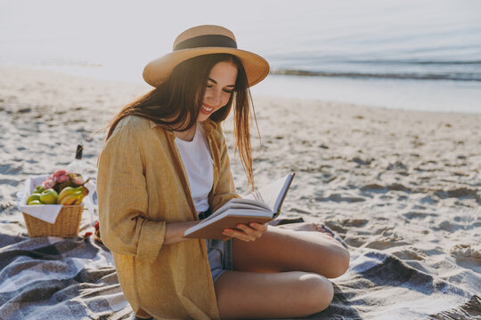 Full length young traveler tourist woman in straw hat glasses shirt summer clothes reading book sit on plaid have picnic outdoor on sea sand beach background People vacation lifestyle journey concept