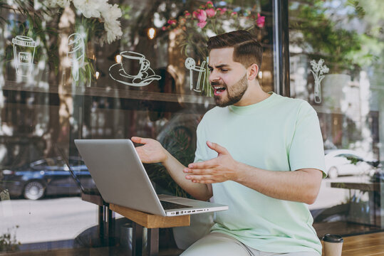 Smiling Freelancer Excited Young Man In Casual Mint T-shirt Work Online With Laptop Pc Computer Use Free Wifi Talk By Video Call Spread Hand Near Outdoors Cafe Urban Leisure Lifestyle Lunch Concept