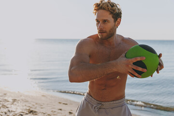 Young sporty athletic toned fit sportsman topless man 20s warm up train play hold medicine crossfit ball at sunrise sun dawn over sea beach outdoor on sand dawn beach seaside in summer day morning.