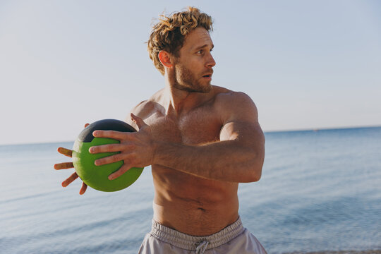 Young sporty athletic toned fit sportsman topless man 20s warm up train play hold medicine crossfit ball at sunrise sun dawn over sea beach outdoor on sand dawn beach seaside in summer day morning.