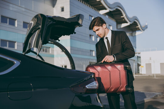 Bottom side view serious young traveler businessman young man in black suit stand outside at international airport terminal put suitcase into car trunk booking taxi Air flight business trip concept