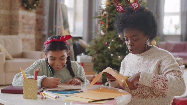 Afro-American girl drawing picture and her mother cutting paper with scissors while making Christmas crafts together at home