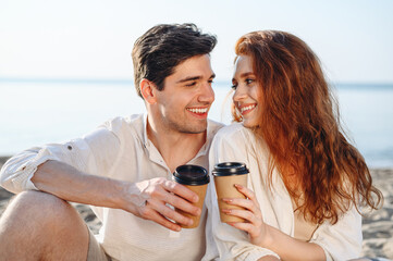 Close up young fun satisfied smiling couple family two man woman in white clothes hug sit on sand have picnic drink coffee rest together at sunrise over sea beach ocean outdoor seaside in summer day