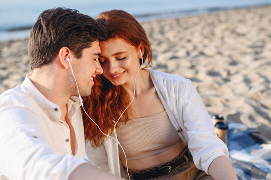 Smiling happy young couple two family man woman in white clothes hug sitting sand listen to music headphones rest together at sunrise over sea beach ocean outdoor seaside in summer day sunset evening.