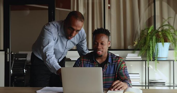 Coaching, apprenticeship concept. African middle-aged mentor give advice, provide professional guidance to young intern colleague using laptop learn software, explain task gather in office workplace