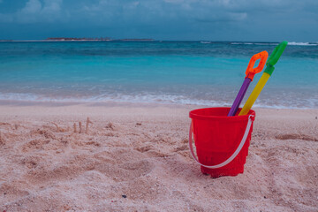 Front view of red toy bucket with shovel inside on the coast of ocean with golden sand and...