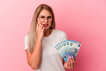 Young russian woman holding bills isolated on pink background biting fingernails, nervous and very anxious.
