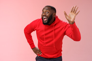 Attractive funny african american gay beard in red hoodie raise palm waving hello hi howdy gesture acting feminine cute fool around welcoming guests greeting standing pink background © Mix and Match Studio
