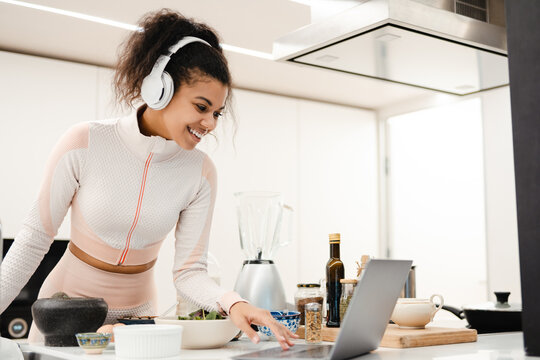 Black woman in headphones using laptop while cooking in kitchen