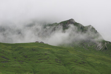 Farm houses, green meadow and mountain peak reaching out of fog and clouds.