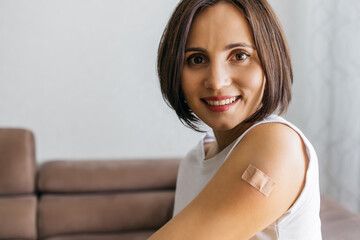 Portrait of smiling woman after getting a vaccine. Female holding down her white shirt sleeve and showing her arm with bandage after receiving vaccination. Concept of recommended inoculation - obrazy, fototapety, plakaty