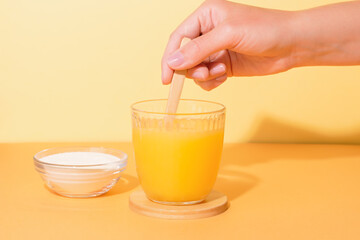 Woman adding collagen powder to orange juice on colorful background