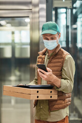 Vertical portrait of delivery man wearing mask and holding pizza while standing in elevator