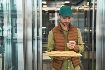 Waist up portrait of delivery man using smartphone while standing by elevator in office building,...