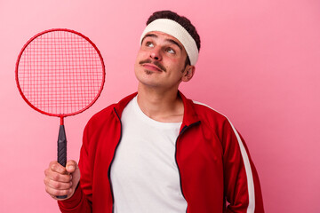 Young caucasian man playing badminton isolated on pink background dreaming of achieving goals and...