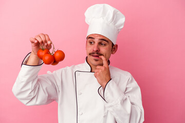 Young caucasian chef man holding tomatoes isolated on pink background relaxed thinking about something looking at a copy space.