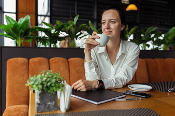 Cheerful businesswoman drinking coffee in cafe while having coffee break during workday and looking away 