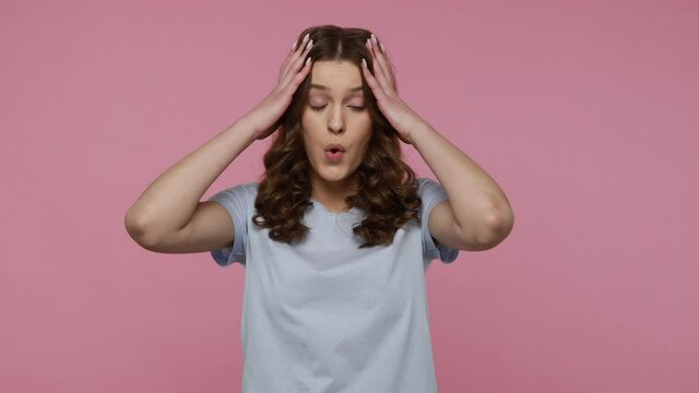 Bored exhausted teenager girl in blue T-shirt touching her head and showing explosion, looking worried and shocked, professional burnout. Indoor studio shot isolated over pink background.