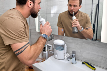 Handsome man brush teeth using electric oral irrigator with jet of water, looking at mirror. Side...