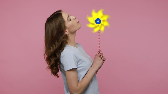 Portrait of happy wavy haired woman in urban style T-shirt looking at camera, smiling carefree and holding windmill, yellow pinwheel toy on stick. Indoor studio shot isolated over pink background.