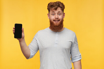 indoor image of young man with big beard, smiles broadly and show display of his phone with a blank black copy space. isolated over yellow background