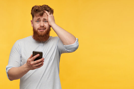 amazed young man with a big red beard staring into camera with a shocked facial expression, holds his phone, get some good news. isolated over yellow background.