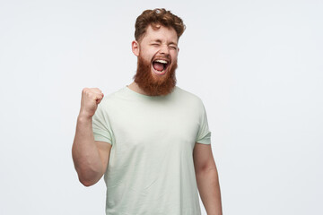 studio shot of young redhead bearded male wears blank t-shirt, raise his fist and yelling while watching football match. victory concept. isolated over white background