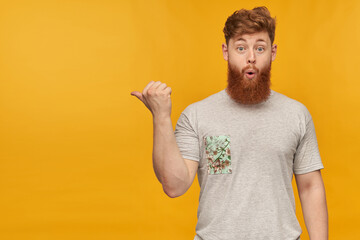 indoor shot of amazed, positive young bearded male with red hair, pointing with a finger left at...