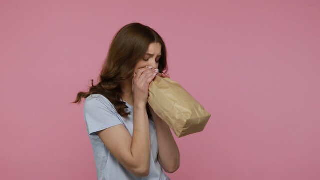 Beautiful wavy haired teenager girl in blue casual T-shirt exhaling and inhaling into package, using paper bag to calm down, overcoming stress fear. Indoor studio shot isolated over pink background.