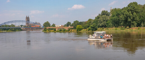 Panoramic view over old cathedral at historical downtown in Magdeburg with a touristic party boat with many girls coming by at Elbe river, Germany, at blue summer sky with clouds.