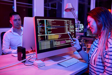 Smiling female software developer sitting at office desk and texting friends instead of coding