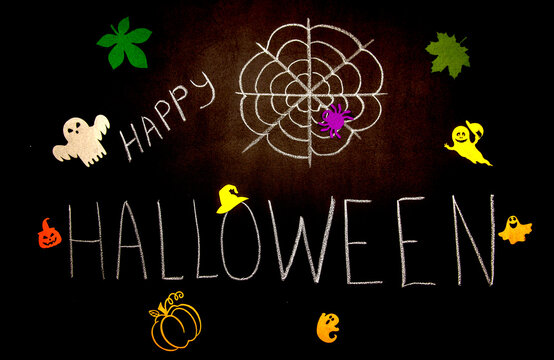The inscription in chalk "Happy Halloween" and a picture of a spider web on a school black chalkboard, little ghosts on a black background, pumpkin and autumn leaves for a scary autumn holiday.
