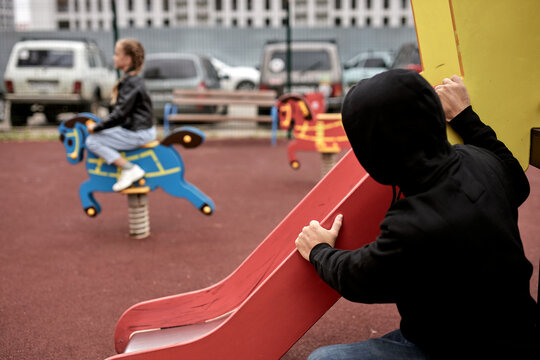 creepy threatening pedophile spies on child girl while having fun in playground alone, in casual wear at day time in city. dangerous man commit a crime, children abuse concept. focus on man