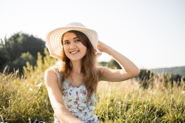 Close up portrait of pretty stunning young woman in dress and hat, sitting on green grass outdoors in the meadow, smiling at camera