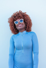 Chica afro, con gafas de sol sonriente, con vestido azul en fondo blanco.