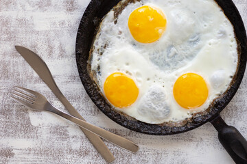 Top view of fried eggs on black pan with fork and spoon on the white background